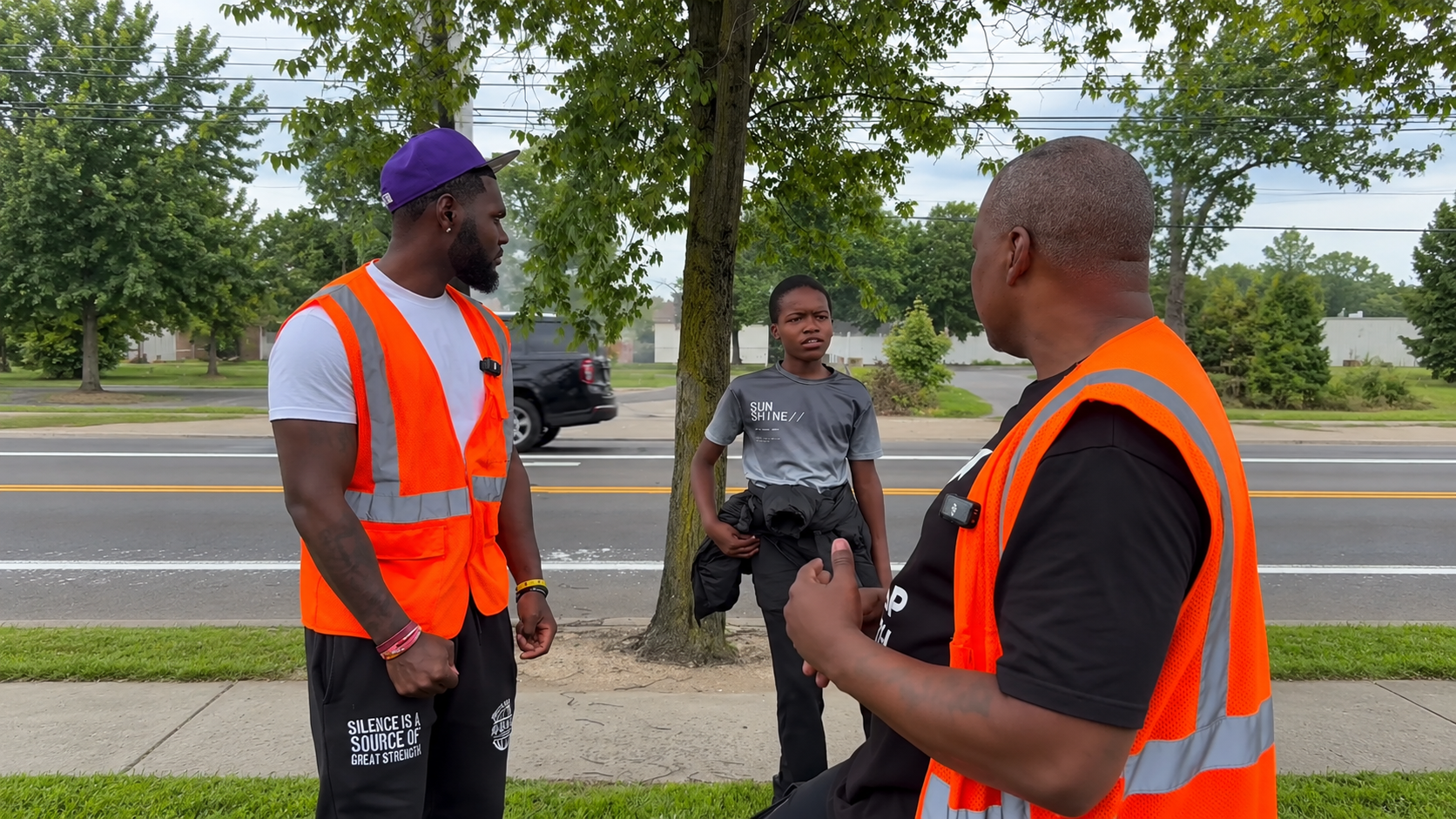 Two Safe Passages team members in orange safety vests talk with a young person along a tree-lined Akron street.